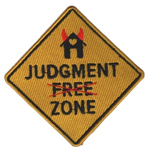 Könnte beinhalten: Ein gelbes, rautenförmiges Schild mit schwarzem Text, der "JUDGMENT FREE ZONE" lautet, wobei das Wort "FREE" durchgestrichen ist. Das Schild zeigt auch ein schwarzes Haus mit einem roten Herzen und roten Teufelshörnern.