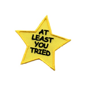 以下が含まれることがあります： 「At Least You Tried」と黒色の刺繍で書かれた黄色い星形のワッペンです。