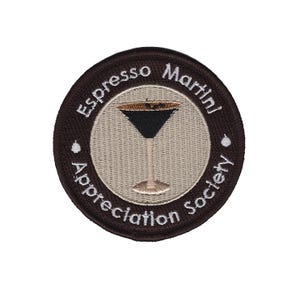 Peut inclure: Écusson brodé circulaire avec les mots "Espresso Martini Appreciation Society" en lettres blanches. L'écusson présente un dessin de verre à martini avec une boisson marron foncé et une tige et une base marron clair, sur fond beige.