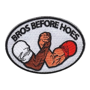 以下が含まれることがあります： 白地に黒文字で「Bros Before Hoes」と刺繍された楕円形のワッペン。2人の人物が腕相撲をしていて、一人は白い腕章、もう一人は赤い腕章をしている。