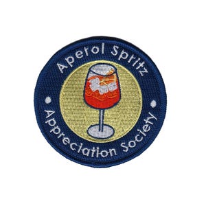 Peut inclure: Écusson brodé rond avec une bordure bleu marine et un centre doré. Le texte "Aperol Spritz Appreciation Society" est brodé sur le bord. Le centre représente un verre à cocktail rempli d'une boisson orange et de glace.