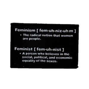 Op de afbeelding: Zwarte patch met witte tekst die de woorden "Feminisme" en "Feministe" definieert met hun fonetische uitspraken en definities. De tekst luidt: "Feminisme [fem-uh-niz-uh m] Het radicale idee dat vrouwen mensen zijn. Feministe [fem-uh-nist] Een persoon die gelooft in de sociale, politieke en economische gelijkheid van de geslachten."