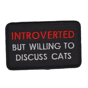Op de afbeelding: Zwarte geborduurde patch met witte en rode tekst die "Introvert maar bereid om over katten te praten" luidt.