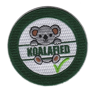 May include: A round green and white embroidered patch with a cartoon koala peeking over a green check mark and the text "KOALAFIED".