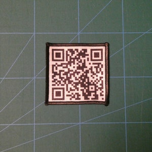 May include: A black and white QR code patch with a black border. The patch is sewn onto a green fabric surface.