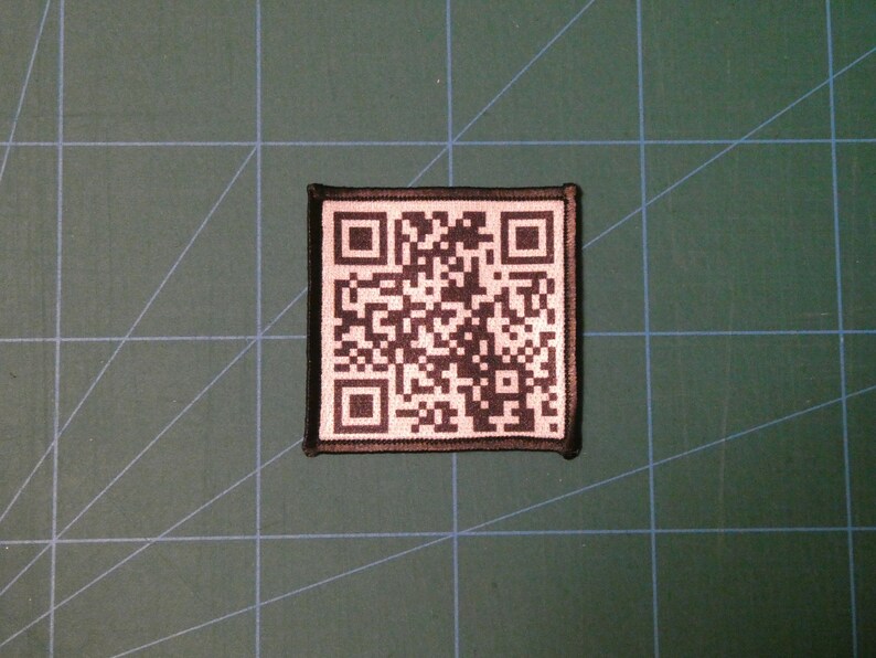 May include: A black and white QR code patch with a black border. The patch is sewn onto a green fabric surface.