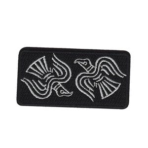 Könnte beinhalten: Ein schwarzer rechteckiger Aufnäher mit zwei stilisierten weißen Vogelmotiven. Die Vögel haben detaillierte Flügel und Körper, die symmetrisch angeordnet sind. Der Aufnäher ist wahrscheinlich für Kleidung oder Accessoires bestimmt.