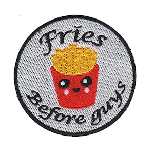 Op de afbeelding: Een ronde, grijs-zwarte geborduurde patch met de tekst "Fries before guys" en een cartoon afbeelding van een rode container met friet met een glimlachend gezicht.