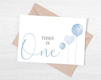 May include: A white card with blue watercolor balloons and the text "Teddy is One" in black script font.
