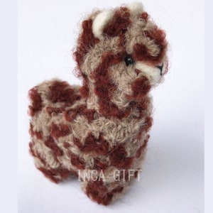 May include: A small, brown and beige alpaca made of wool felt. The alpaca has a white face and black eyes. The alpaca is standing on a white surface. INCA GIFT is printed on the bottom of the alpaca.