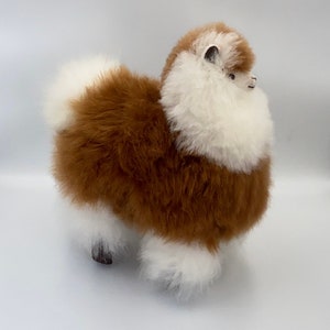 May include: A soft alpaca toy, featuring a brown and white fur design. The plush animal has a fluffy texture, with a brown body and white detailing on the head, tail, and legs. The alpaca has small black eyes and ears.
