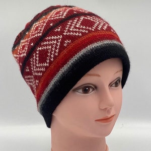 May include: A knitted beanie hat with a geometric pattern. The hat features a color scheme of red, black, white, and orange. The design includes diamond shapes and stripes. The brim is black, and the hat is displayed on a mannequin head.