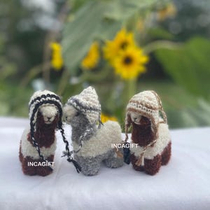 May include: Three small alpaca plushies wearing knitted hats. The alpaca on the left is brown and white, the middle alpaca is gray and white, and the alpaca on the right is brown and white. The plushies are all wearing hats with different patterns.