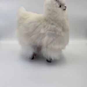 May include: A white alpaca ornament with a fluffy, textured coat and small, dark ears. The alpaca has black eyes and stands on four dark legs. The figurine is set against a plain white background, highlighting its soft appearance.