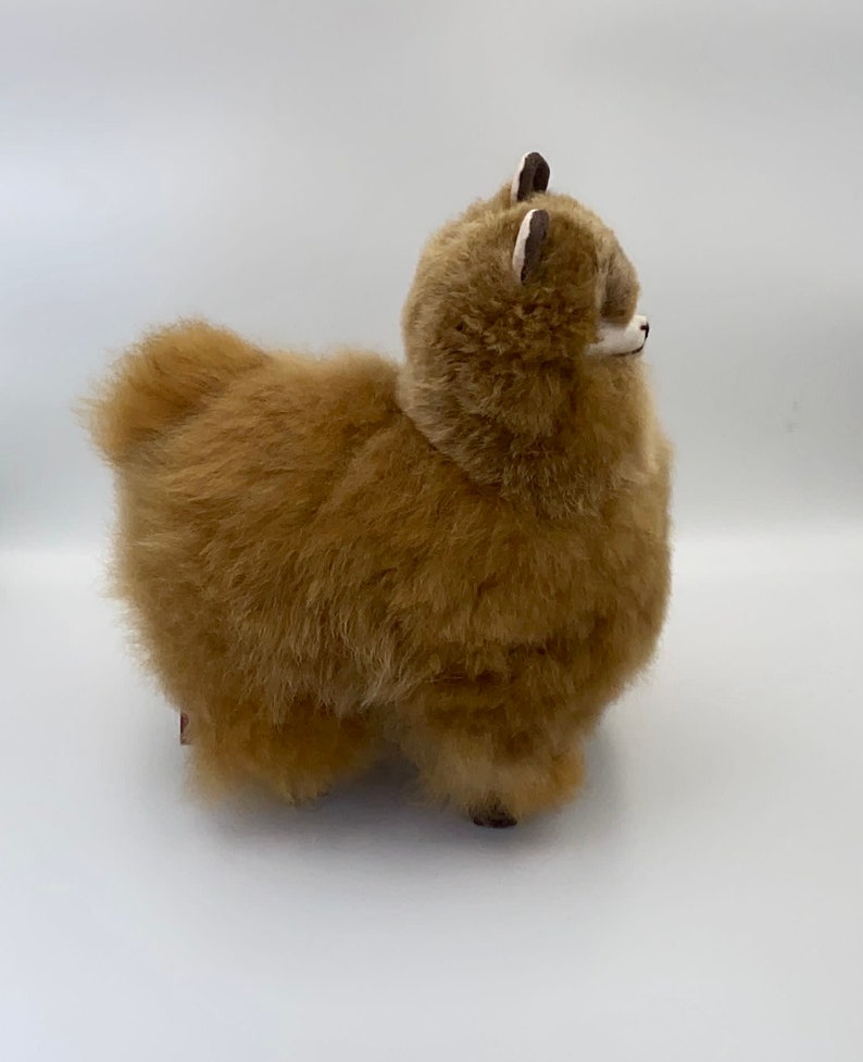 May include: A soft, brown alpaca toy with a fluffy coat. The alpaca has small, dark ears with white detailing and a white face. The figurine stands on four small legs, and the overall design is soft and cuddly, perfect for a child's room.