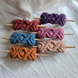 May include: A collection of six macrame hair accessories in purple, rust, blue, pink, peach, and orange. Each piece features a knotted cord design wrapped around a smooth, pointed wooden stick, creating a textured, cylindrical hair ornament.
