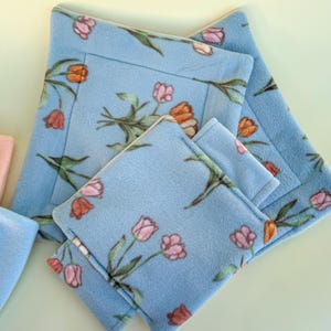 May include: Three blue fleece squares with a floral pattern of pink and orange tulips. The squares are nested inside each other, with the largest square on the bottom and the smallest square on top.