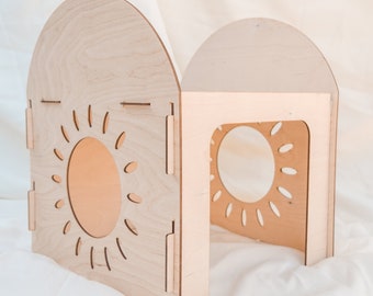 May include: A wooden playhouse for children, featuring a sun-shaped cutout on each side. The playhouse is made of light-colored wood and has a rounded roof.