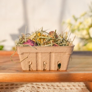May include: A small, brown, berry basket filled with dried herbs, grasses, and flower petals. The basket sits on a wooden surface, with a blurred background of flowers and sunlight.