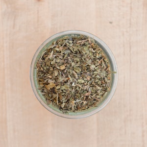 May include: A glass jar filled with dried green herbs, likely mint, on a light wood surface.