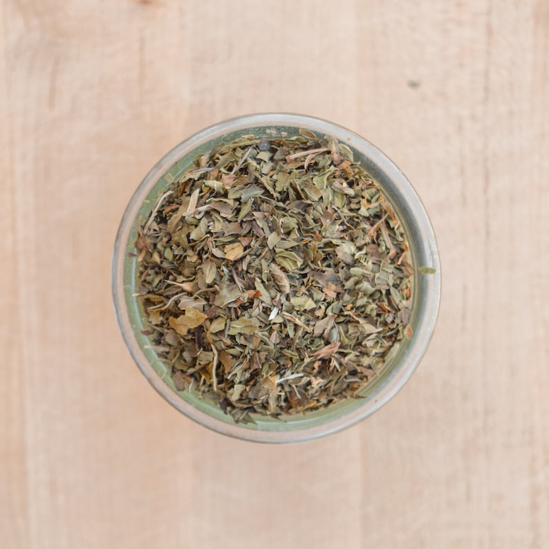 May include: A glass jar filled with dried green herbs, likely mint, on a light wood surface.