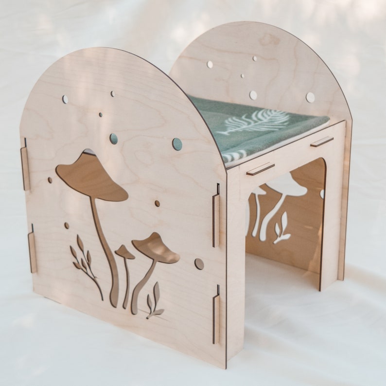 Puede incluir: Una casa de madera para gatos con forma de hongo con un coj&iacute;n verde en el interior. La casa tiene un dise&ntilde;o recortado de hongos en la parte delantera y los lados.