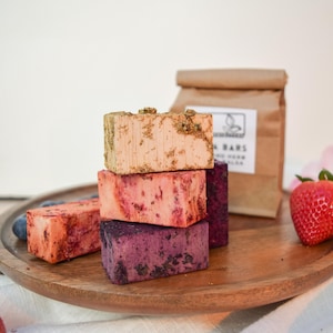 May include: A stack of four colorful fruit and herb bath bars. The bars are arranged in a pyramid shape, with the top bar being a light brown color with gold flecks, the second bar is a pink color with red streaks, the third bar is a dark purple color with black flecks, and the bottom bar is a light orange color. The bars are sitting on a wooden surface.