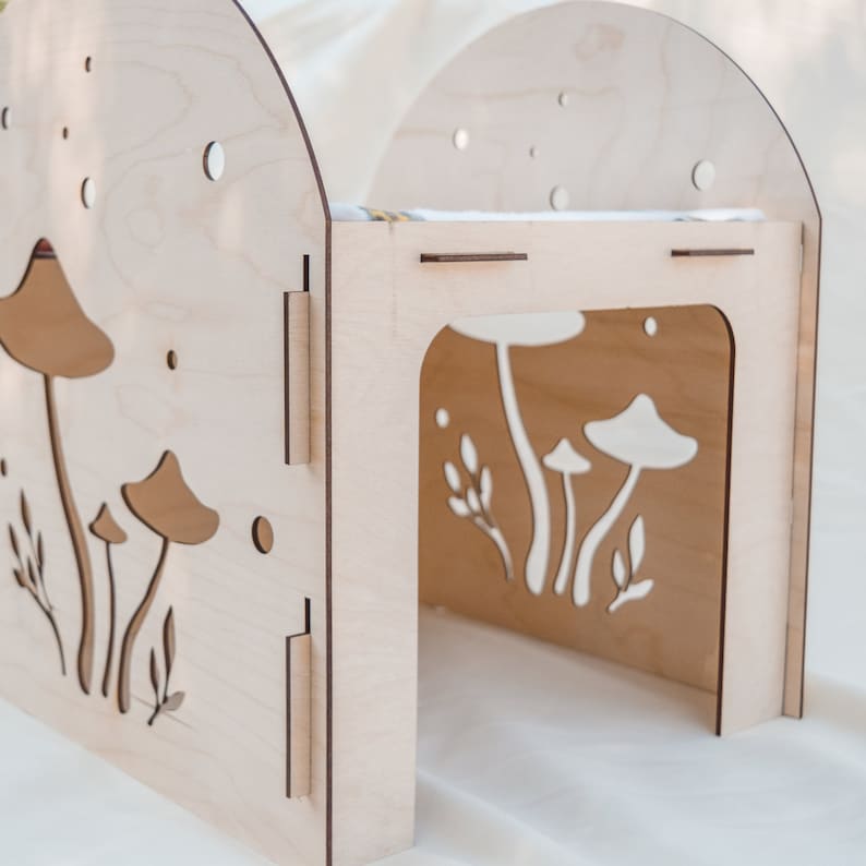 Puede incluir: Una casa de madera para gatos con forma de hongo con un dise&ntilde;o recortado de hongos y hojas. La casa est&aacute; pintada en un color madera claro y tiene una peque&ntilde;a abertura para que el gato pueda entrar.