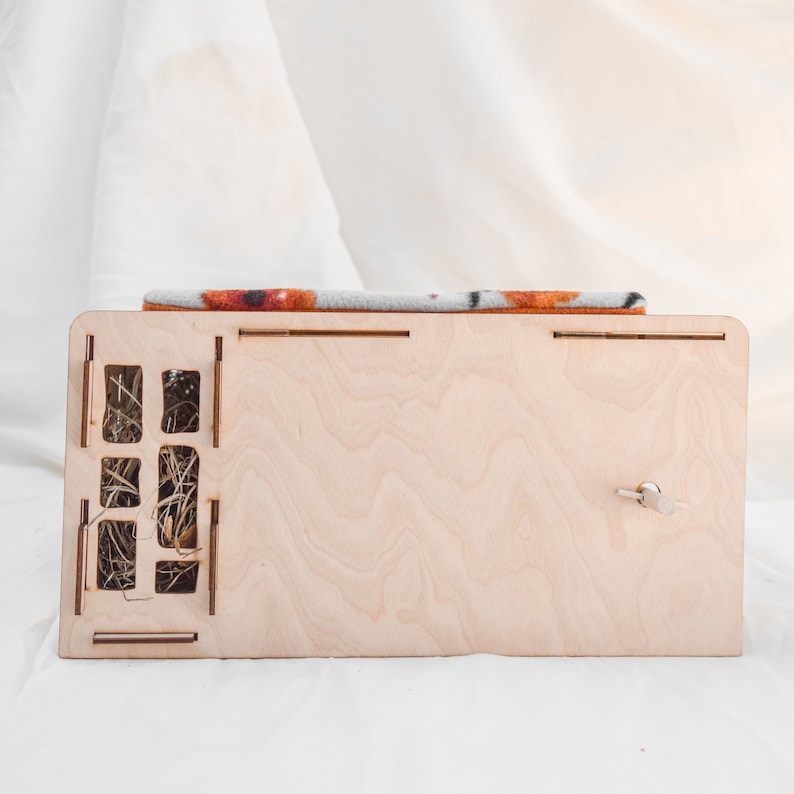 May include: A wooden pet house with a removable top and a small door. The house is made of light wood and has a brown and gray patterned fabric lining. The house is sitting on a white surface.