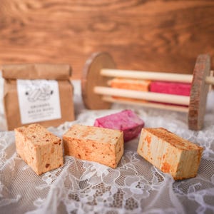 May include: Three rectangular orange-speckled blocks and one pink block arranged on white lace fabric. A brown paper bag with a white label featuring black text and illustrations sits behind the blocks, alongside a wooden rack with dowels.