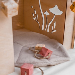 Puede incluir: Una casa de juegos de madera con un coj&iacute;n gris suave en el interior. La casa de juegos tiene un dise&ntilde;o recortado de hongos y hojas en la pared posterior. Hay tres bloques de madera con una cuerda atada, dos son beige y uno es rojo.