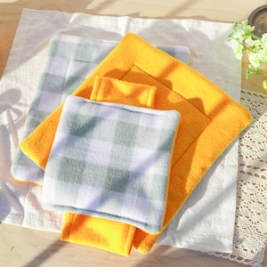 May include: A collection of pet bed pads in various sizes and colors. The pads include a vibrant yellow fleece pad, and pads with a gray and white checkered pattern. The pads are arranged on a white cloth.