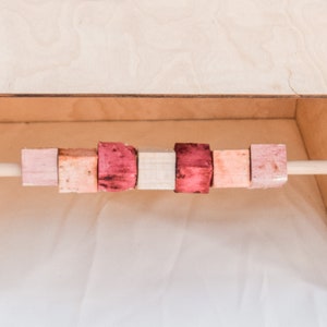 May include: A wooden dowel rod with six colorful wooden blocks strung on it. The blocks are pink, light brown, dark pink, light brown, light pink, and dark pink.