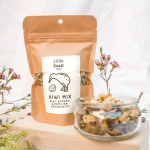 May include: A brown paper bag of "Kiwi Mix" pet treats with a kiwi bird logo, and a small glass jar filled with treats. The bag is labeled with the ingredients: kiwi, banana, dandelion, and meadowsweet. Pink flowers and greenery are in the background.