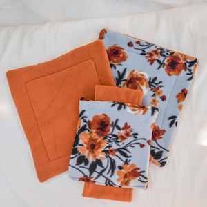 May include: Three fleece fabric squares with floral patterns. Two squares are blue with orange and brown floral designs. One square is solid orange.