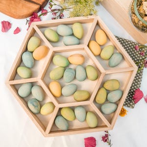 May include: A wooden, geometric tray filled with pastel-colored, oval-shaped wooden toys. The toys are in shades of yellow, green, and blue. The tray is made of light-colored wood and has a hexagonal shape with internal dividers.