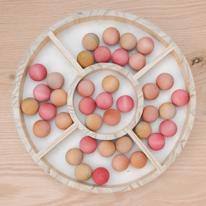 May include: A wooden tray with a circular design, divided into sections. The tray holds numerous wooden balls in shades of pink, peach, and natural wood tones. The balls are arranged within the tray's compartments, creating a visually appealing pattern.