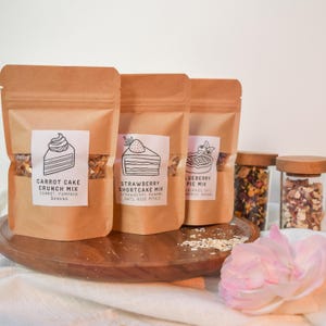 May include: Three brown paper bags with labels and text that reads "Carrot Cake Crunch Mix", "Strawberry Shortcake Mix", and "Blueberry Pie Mix". The bags are filled with a mixture of oats, dried fruit, and other ingredients. Two glass jars with lids are also visible, one filled with dried flowers and the other with a mixture of oats and dried fruit.