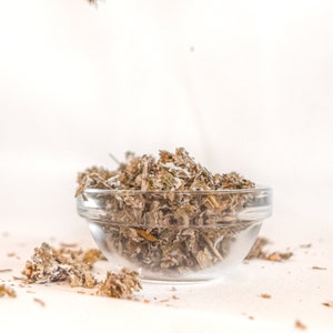 May include: A clear glass bowl filled with dried herbs, with some herbs scattered on a white surface.