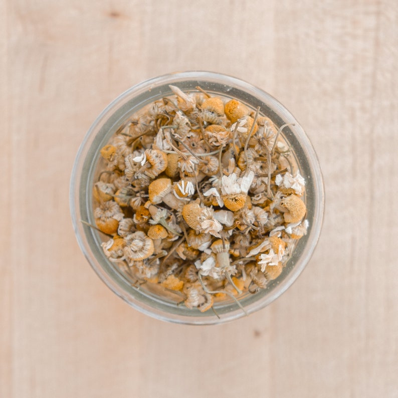 May include: A glass jar filled with dried chamomile flowers. The flowers are a light yellow color and have a delicate, dried texture.