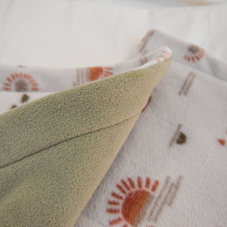 Puede incluir: Primer plano de una tela de franela suave de color verde doblada sobre una tela blanca con un patr&oacute;n repetitivo de dise&ntilde;os de soles naranja y marr&oacute;n.
