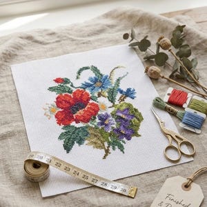 May include: A cross-stitch embroidery piece on white fabric featuring a floral bouquet with red, blue, and purple flowers and green leaves. A yellow measuring tape shows measurements up to 30 cm, alongside gold scissors, embroidery floss, and a tag.
