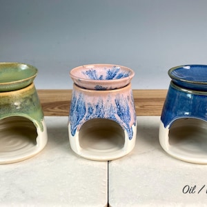 Könnte beinhalten: Drei Öl-/Wachs-Brenner aus Keramik. Die Brenner haben eine abgerundete Oberseite und eine gebogene Öffnung. Die Farben sind Grün, Rosa und Blau, mit weißen Basen. Der Text "Oil / Wax Burner" steht unten.