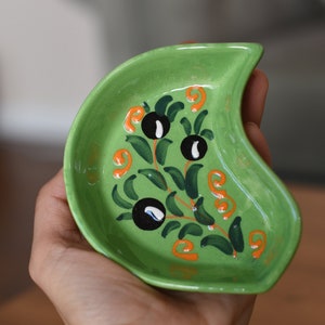 May include: A green ceramic dish with a crescent shape and a hand-painted design of olive branches with black olives and orange swirls.