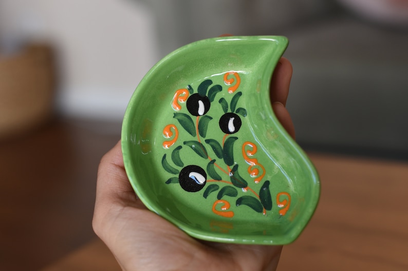 May include: A green ceramic dish with a crescent shape and a hand-painted design of olive branches with black olives and orange swirls.