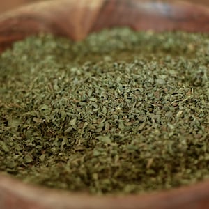 May include: A wooden bowl filled with dried green herbs. The herbs are finely chopped and have a slightly textured appearance. The bowl is made of a light brown wood. The image is a close-up.