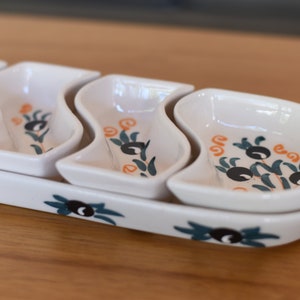 May include: White ceramic serving dish with four individual bowls. The bowls are shaped like a wave and have a green and orange olive branch design.