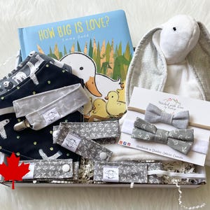 May include: A gift box filled with baby items, including a book titled "How Big Is Love?" by Emma Dodd, a grey and white bunny plush toy, a grey and white bow tie, and a grey and white bib with a black and white pattern. The box is filled with shredded paper and has a Canadian flag sticker on the side.