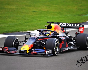 Puede incluir: Un coche de carreras de Fórmula 1 negro y rojo con las palabras "Red Bull" y "Honda" en el lateral. El coche está en una pista de carreras con hierba verde en el fondo.