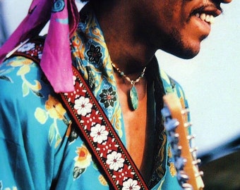 May include: A man wearing a blue and floral patterned shirt with a pink bandana and a guitar strap with white flowers and red accents. He is playing a guitar with a light brown body and a silver headstock.